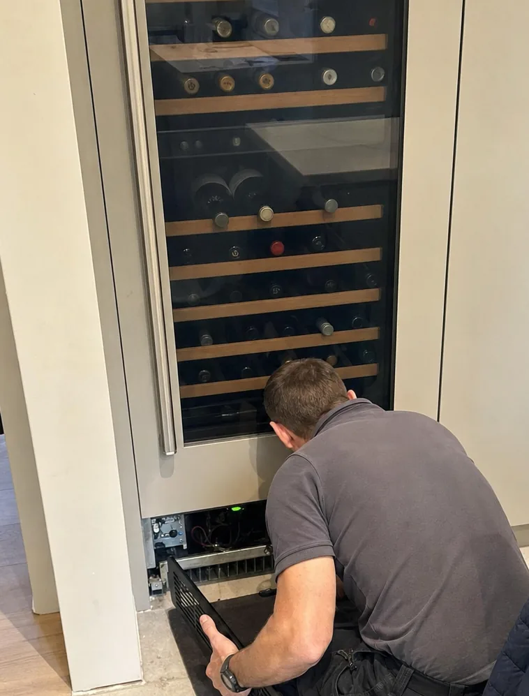 A wine column photographed from the middle of the room, a technician working low at its base with the grille off.